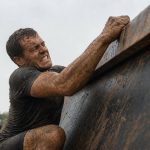 A mud-covered obstacle racer straining to haul himself over a steep inverted wall on an off-road course