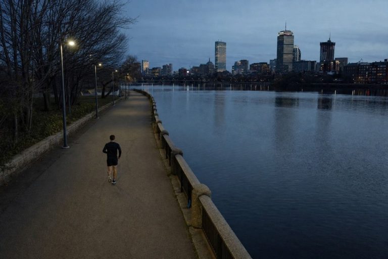 A runner on a riverside path at dusk with the city lights coming on behind